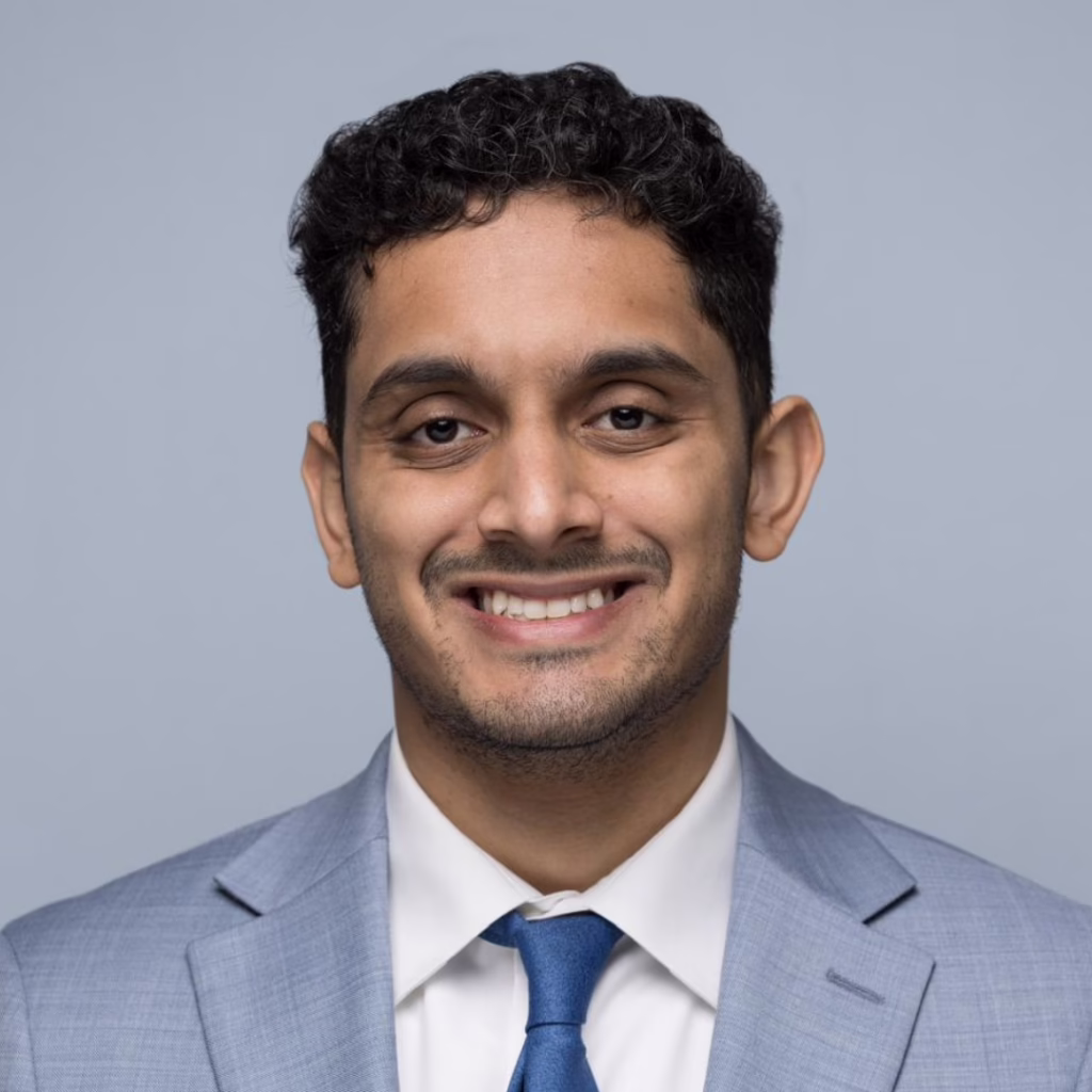 Head-and-shoulders portrait of a smiling man in a light blue suit and blue tie against a gray background.